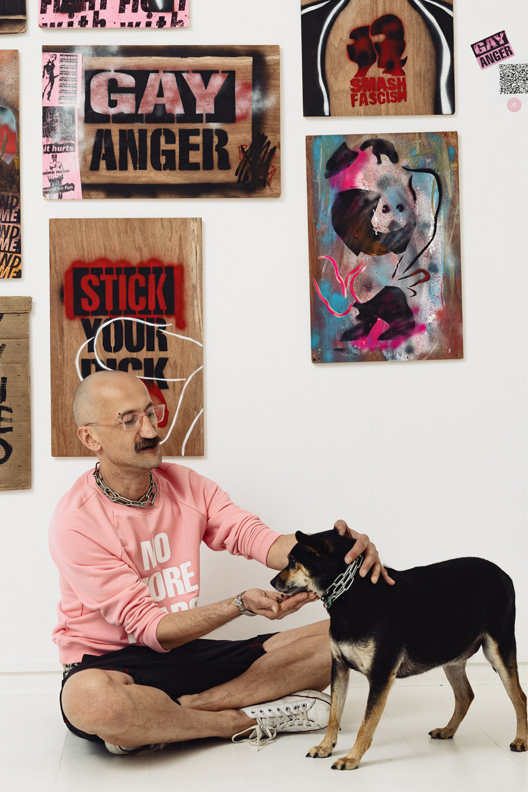 Mężczyzna w różowej bluzie z napisem 'NO MORE' głaszcze psa na tle sztuki ulicznej z napisami 'GAY ANGER' i 'SMASH FASCISM'