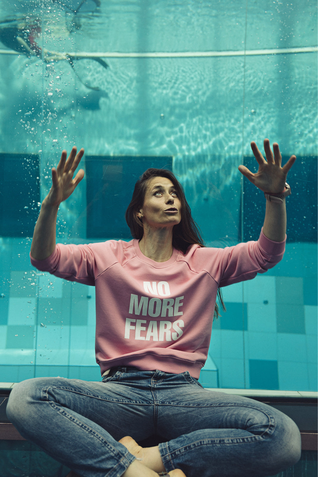 Różowy sweter z napisem NO MORE FEARS, jeansy, kobieta pod wodą w basenie – moda self-care, precyzyjne krawiectwo