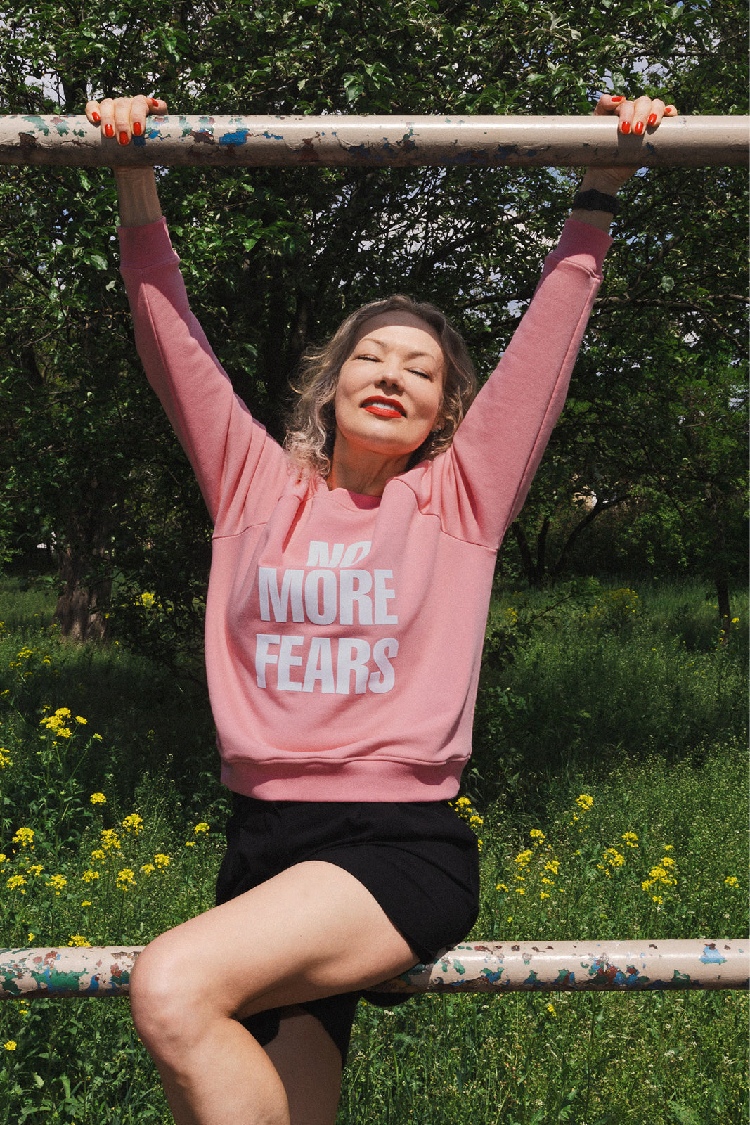Różowy sweter z napisem 'NO MORE FEARS', kobieta na barierce w zielonym parku, modna stylizacja