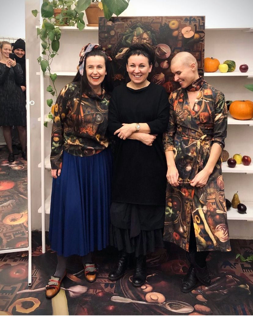 Trzy kobiety w stylowych sukienkach i bluzach, uśmiechnięte w eleganckim wnętrzu z owocami i roślinami.