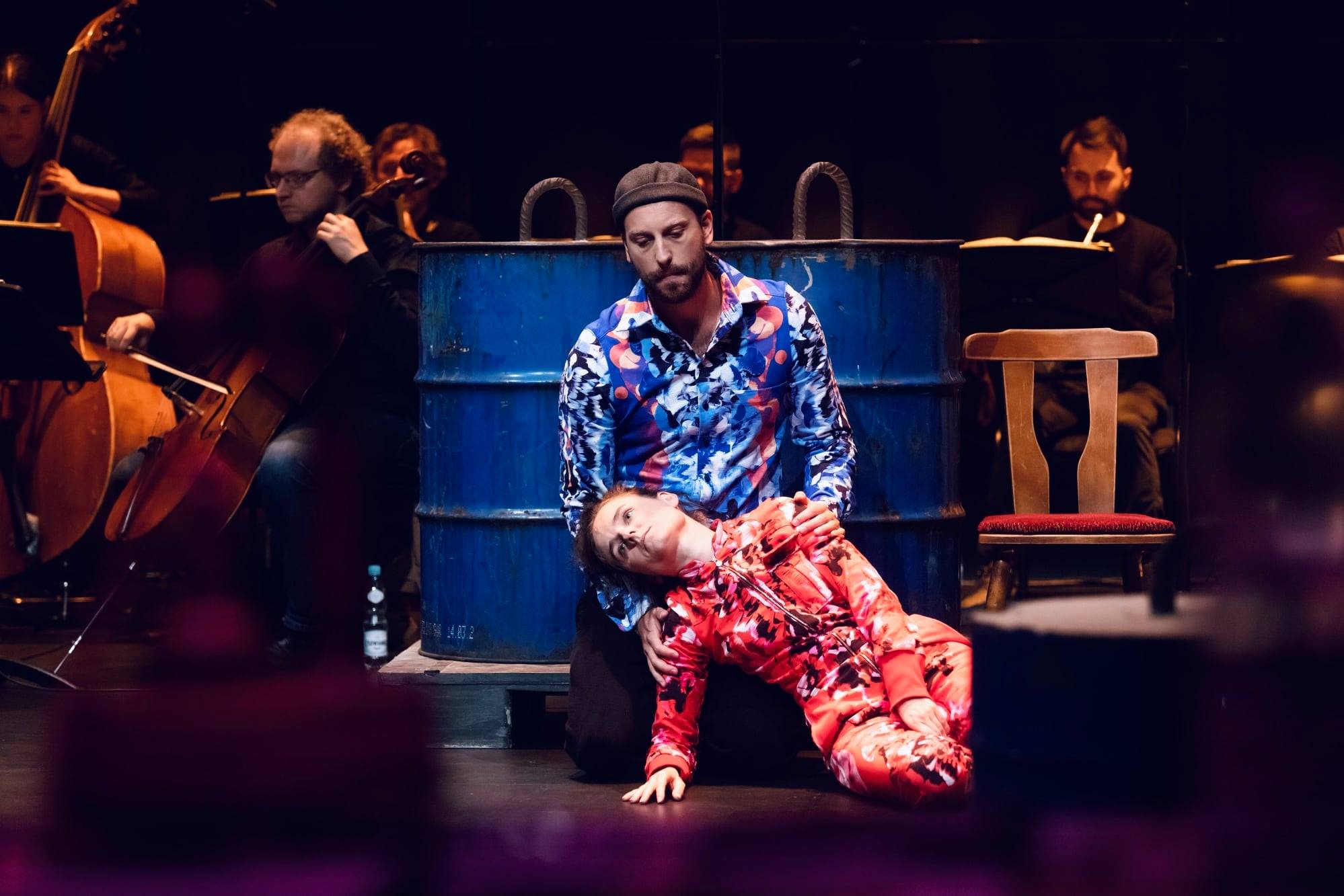 Kobieta w czerwonej bluzie i spodniach w kwiaty, wspierana przez mężczyznę w kolorowej koszuli na scenie z muzykami.